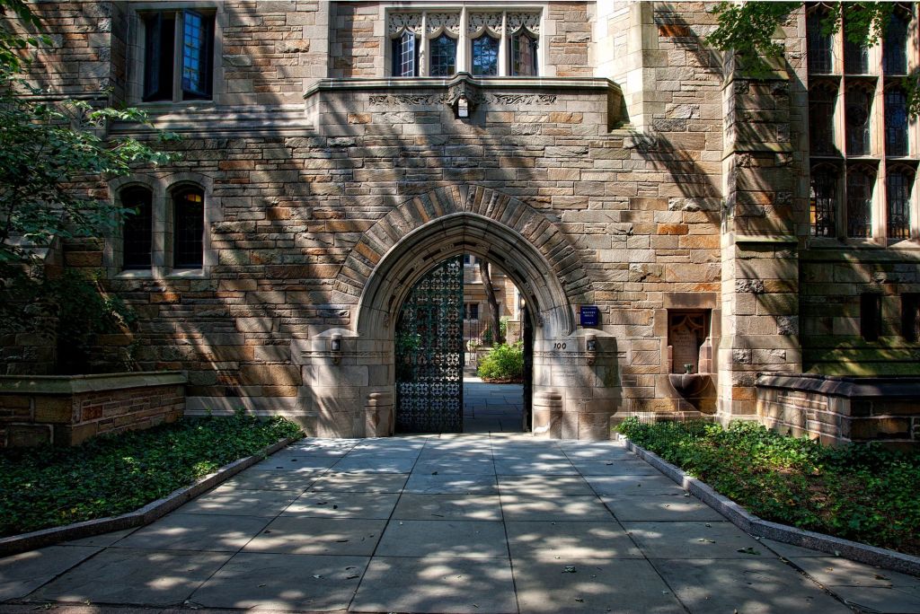 A stone archway entrance flanked by greenery, leading into a historic building with intricate architecture and shadows cast on the pathway.